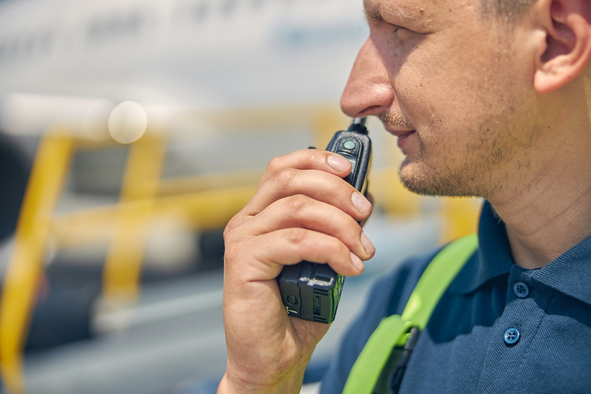 A close-up side profile of a man wearing a dark blue polo shirt and a bright green safety harness strap, holding a black walkie-talkient.
